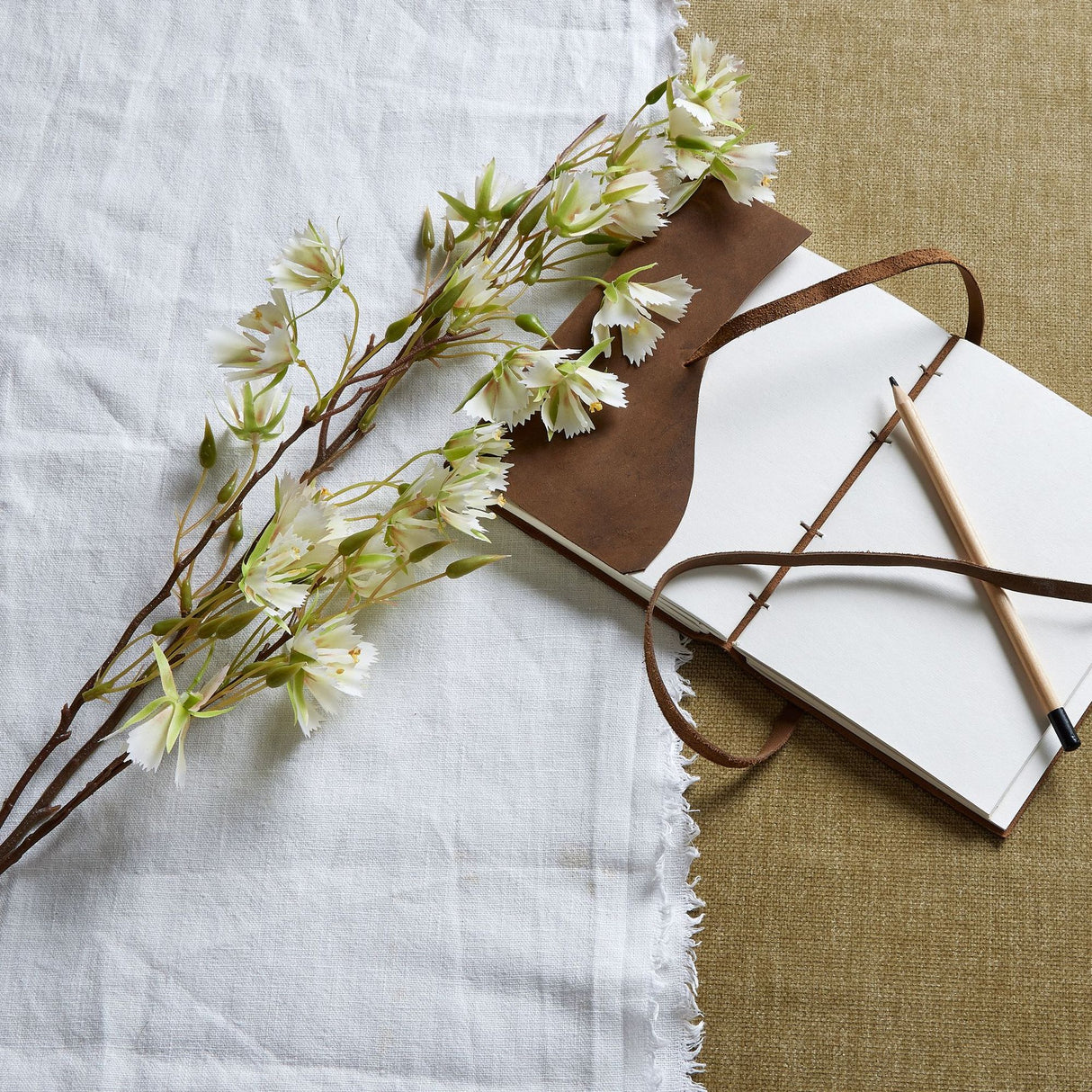 Hill Interiors Elaeocarpus Stem faux flower branch with white blooms and green foliage laid on a white linen cloth beside an open leather journal and pencil.