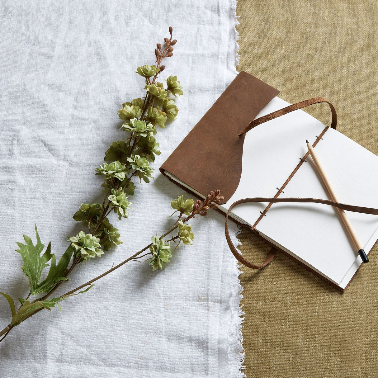 Hill Interiors Tall Green Hops Stem laid on white linen beside a leather-bound journal with pencil on a neutral hessian background.