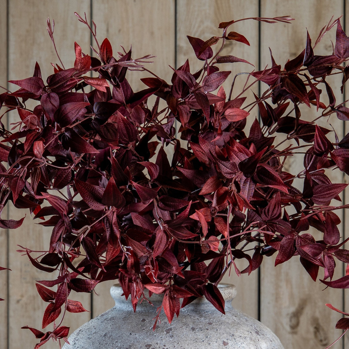 Hill Interiors Ornamental Burgundy Leaf faux foliage stem with deep red veined leaves, arranged in a stone ceramic vase against a light wood plank backdrop.