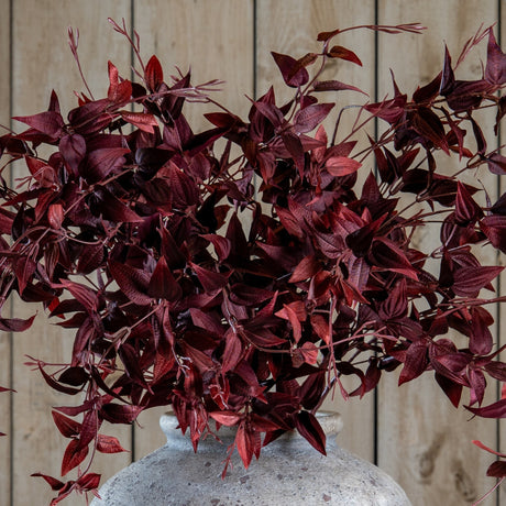 Hill Interiors Ornamental Burgundy Leaf faux foliage stem with deep red veined leaves, arranged in a stone ceramic vase against a light wood plank backdrop.