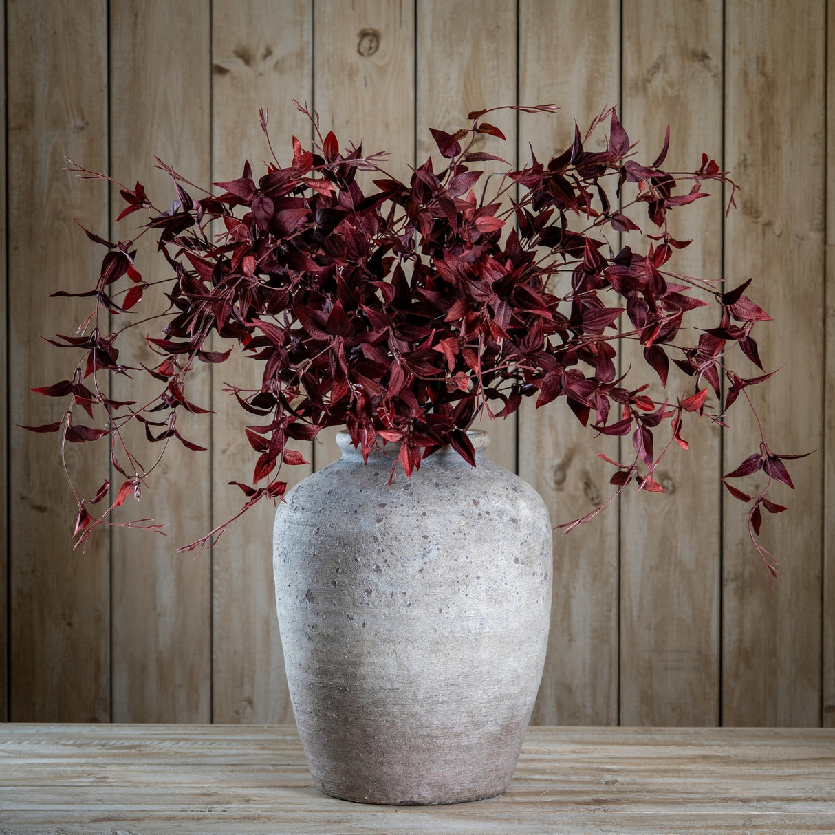 Hill Interiors Ornamental Burgundy Leaf faux foliage stems arranged in a rustic stone pot against a wooden plank backdrop.