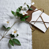 Hill Interiors White Florida Clematis faux flower stem with open blooms, buds and green leaves, laid on white linen beside a leather journal and pencil.