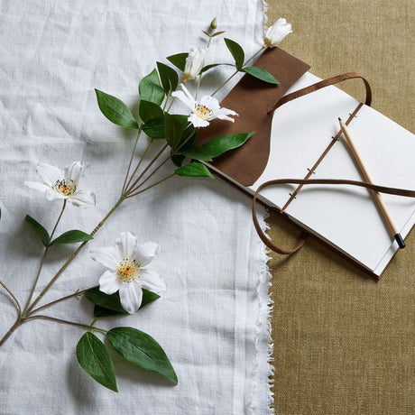 Hill Interiors White Florida Clematis faux flower stem with open blooms, buds and green leaves, laid on white linen beside a leather journal and pencil.