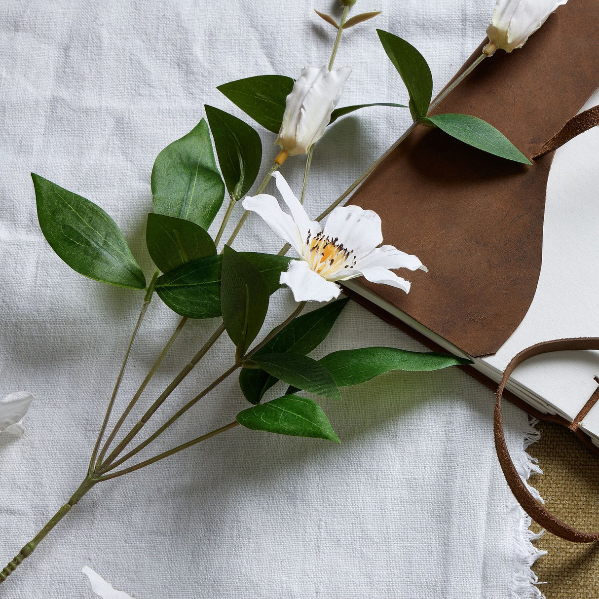 Hill Interiors White Florida Clematis faux flower stem with open bloom, buds and green leaves, laid on linen beside an open leather-bound journal.