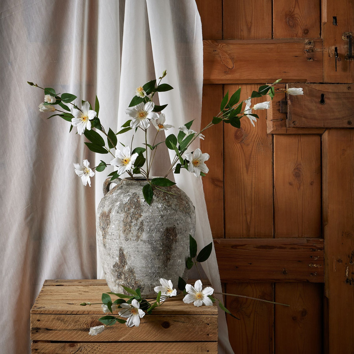 Hill Interiors White Florida Clematis faux stems with layered white blooms and green foliage arranged in a rustic stone urn on a wooden crate.