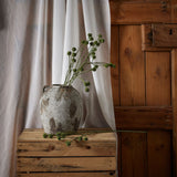 Hill Interiors Green Craspedia Stems arranged in a weathered stone urn on a wooden crate, with sheer curtain and rustic wood panel backdrop.