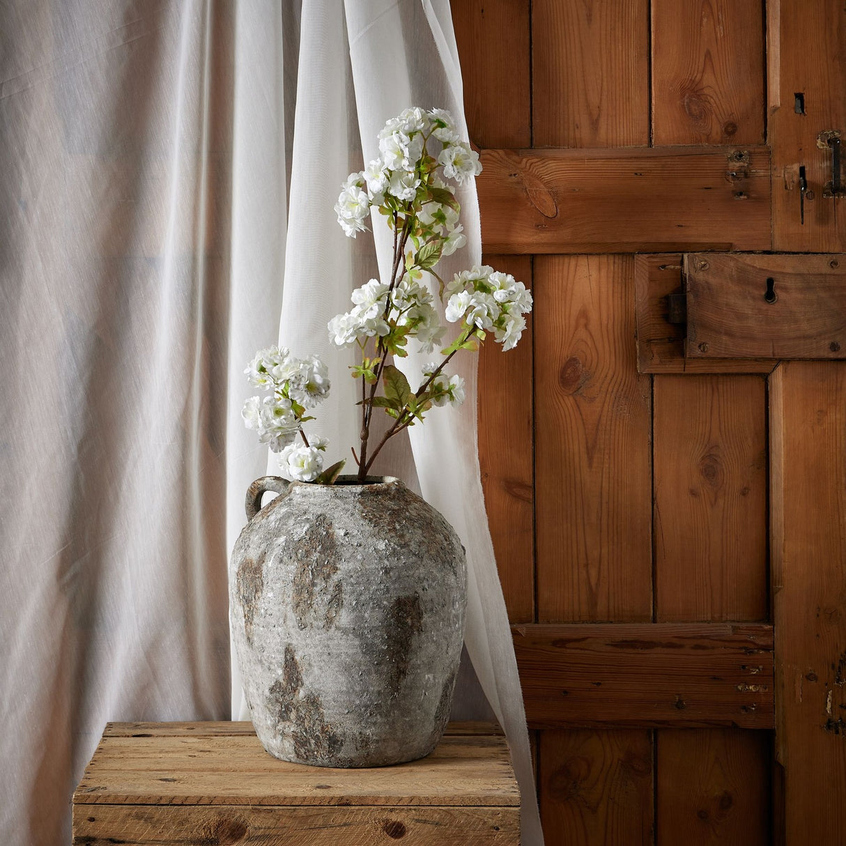 Hill Interiors Tall White Blossom faux stems arranged in a weathered stone vase on a wooden crate beside sheer curtains and a rustic wooden door.
