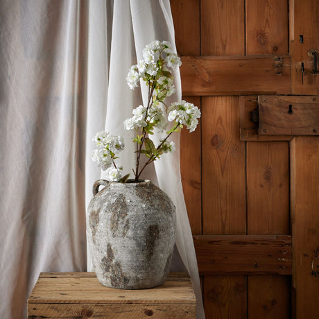Hill Interiors Tall White Blossom faux stems arranged in a weathered stone vase on a wooden crate beside sheer curtains and a rustic wooden door.