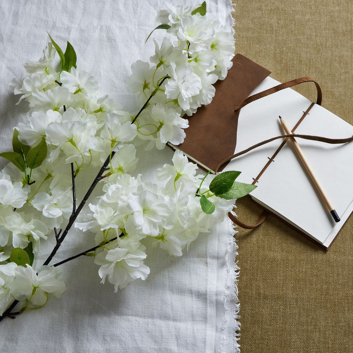 Hill Interiors large white full cherry blossom faux stem laid on white linen beside a leather-bound notebook and pencil on hessian.
