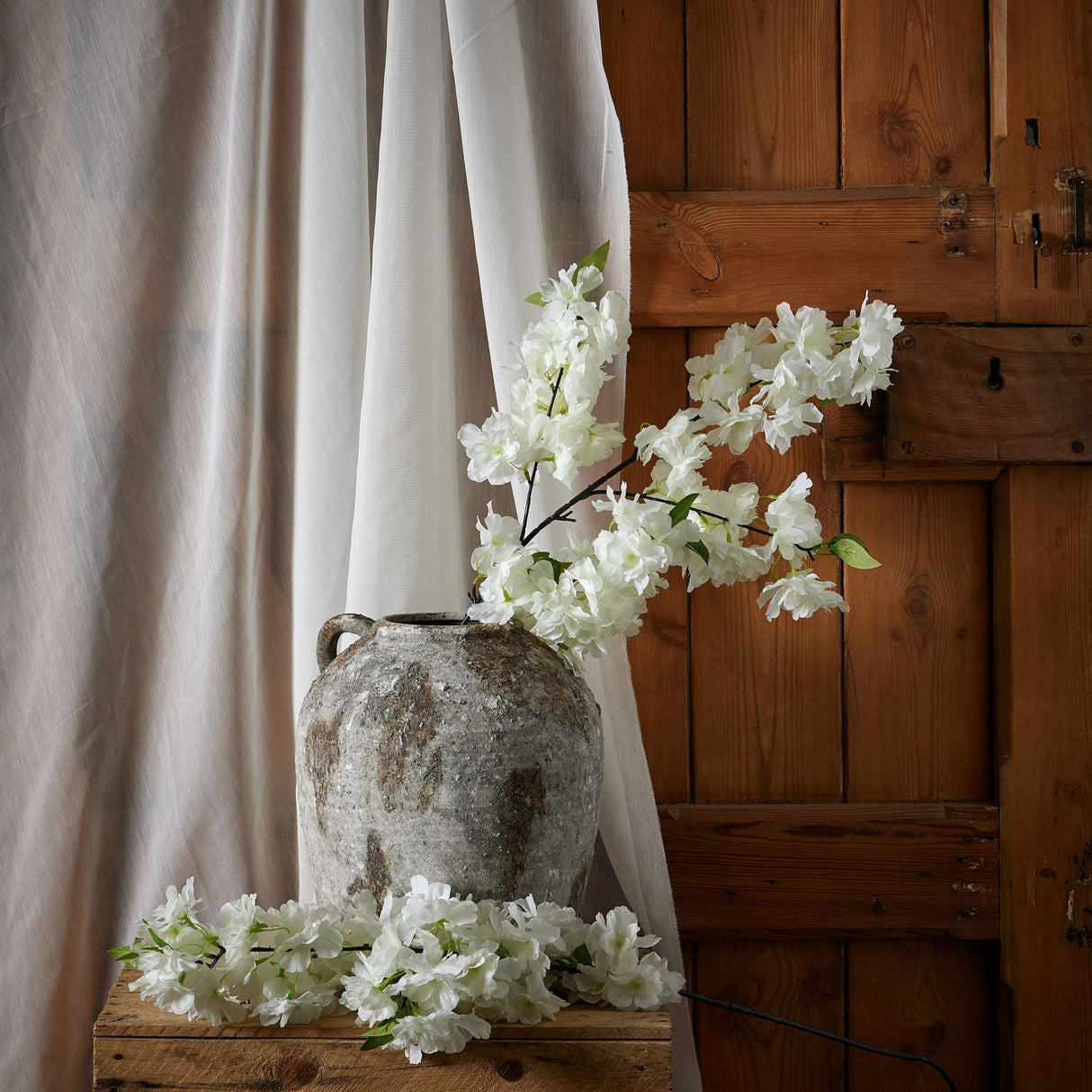 Hill Interiors Large White Full Cherry Blossom Stem arranged in a rustic stone urn on a wooden bench beside sheer curtains and a timber door.