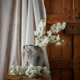 Hill Interiors Large White Full Cherry Blossom Stem arranged in a rustic stone urn on a wooden bench beside sheer curtains and a timber door.