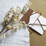 Hill Interiors White Japanese Blossom faux stem with delicate white flowers, laid on white linen beside a leather-bound sketchbook and pencil.