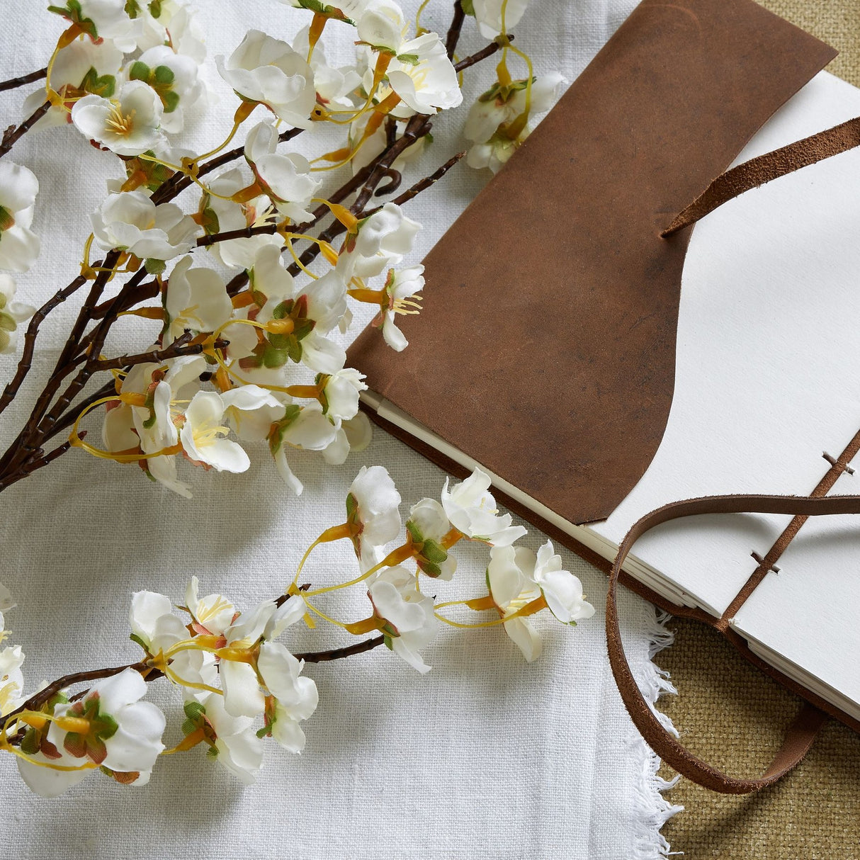 Hill Interiors White Japanese Blossom faux stem with clusters of white petals and slim buds, laid beside a brown leather-bound journal on linen.