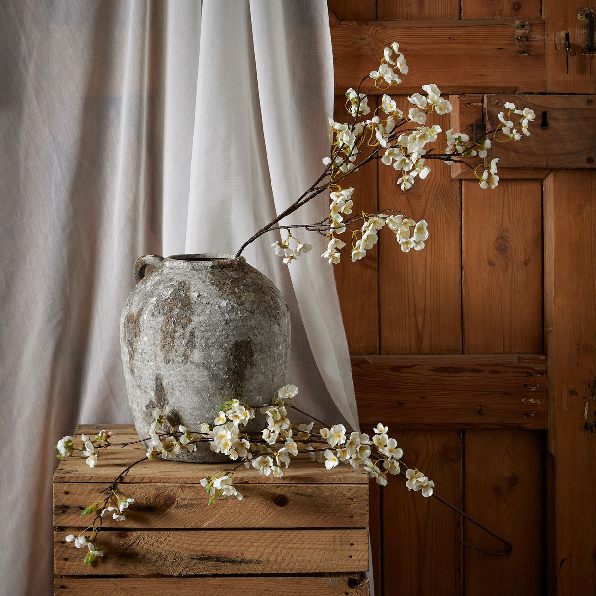 Hill Interiors White Japanese Blossom faux stems arranged in a weathered stone vase on a rustic wooden crate beside linen drapery and a timber door.