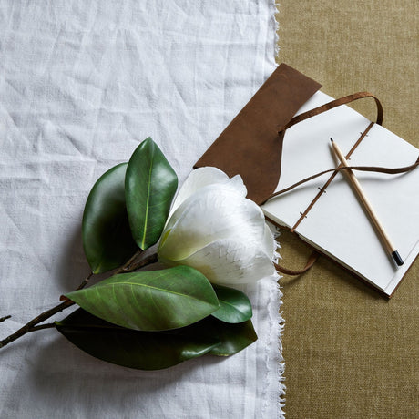 Hill Interiors White Magnolia Stem with waxy petals and green leaves resting on linen beside a leather-bound notebook and pencil.