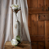 Hill Interiors White Magnolia Stem displayed in a tall weathered stone bottle vase with a second bloom resting on a wooden crate beside draped linen.