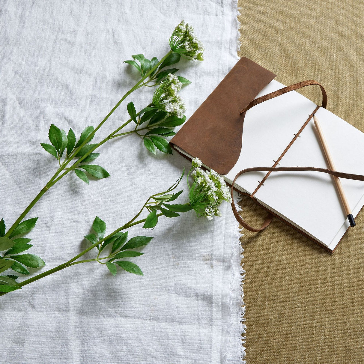 Hill Interiors Faux White Cow Parsley Ammi stems with delicate white florets and green foliage, laid on white linen beside a leather-bound journal and pencil.