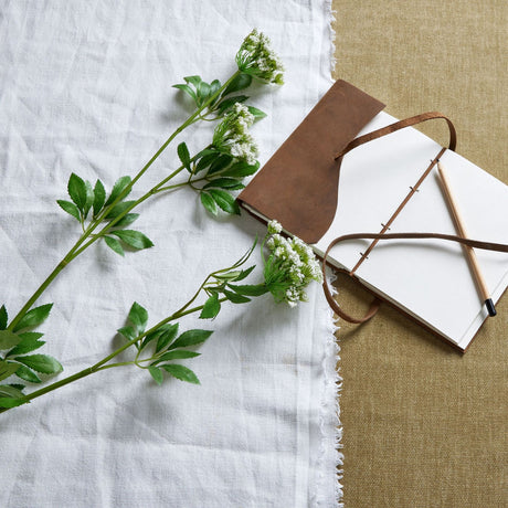 Hill Interiors Faux White Cow Parsley Ammi stems with delicate white florets and green foliage, laid on white linen beside a leather-bound journal and pencil.