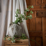 Hill Interiors Faux White Cow Parsley Ammi stems arranged in a weathered stone urn on a wooden crate against a linen curtain and rustic wooden door.