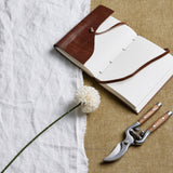 Hill Interiors White Short Chrysanthemum faux stem laid beside an open leather-bound journal and wooden-handled secateurs on linen and hessian.