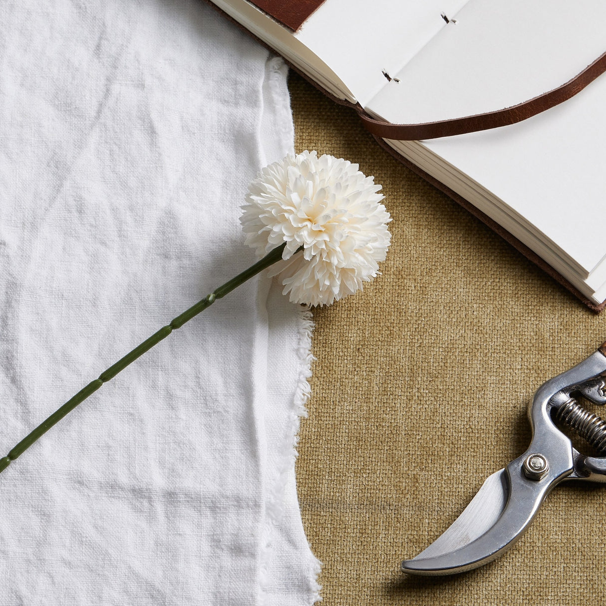 Hill Interiors White Short Chrysanthemum faux stem with domed layered petals, laid on white linen beside an open leather journal and steel secateurs.