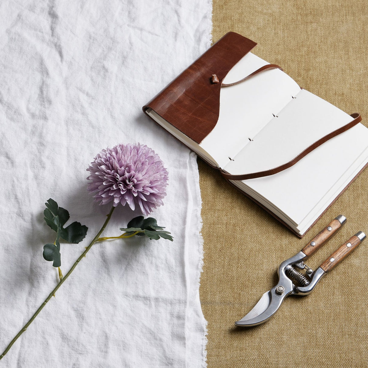 Hill Interiors light purple chrysanthemum faux stem with layered petals and green leaves on white linen beside a leather journal and wooden pruning shears.
