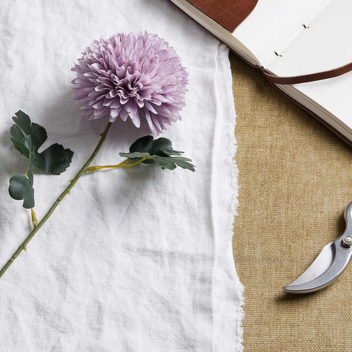 Hill Interiors Light Purple Chrysanthemum faux stem with layered pom-pom bloom and green leaves on white linen beside an open leather journal and secateurs.