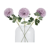 Three Hill Interiors light purple chrysanthemum faux stems with pom-pom blooms and green leaves arranged in a clear glass vase on a white background.