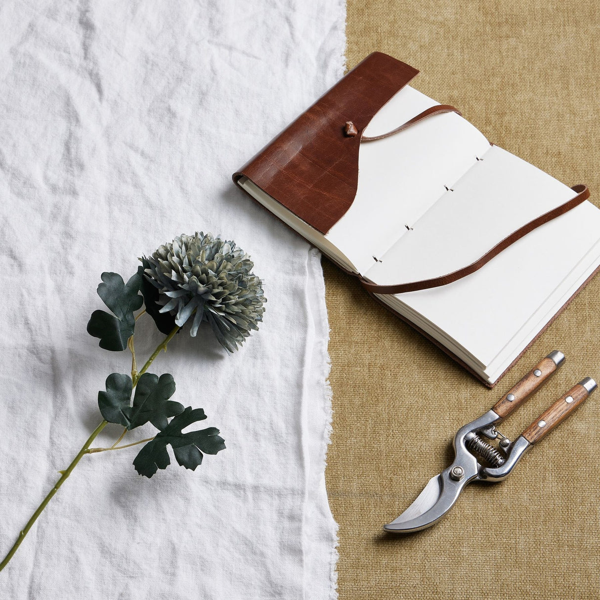 Hill Interiors Pale Green Blue Chrysanthemum faux stem laid on white linen beside a leather journal and wooden-handled secateurs on hessian.