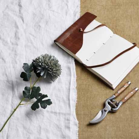 Hill Interiors Pale Green Blue Chrysanthemum faux stem laid on white linen beside a leather journal and wooden-handled secateurs on hessian.