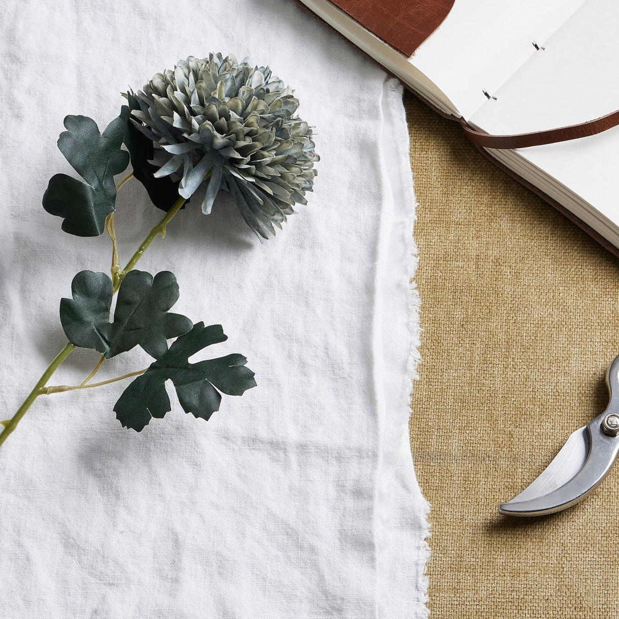 Hill Interiors Pale Green Blue Chrysanthemum faux stem with layered petals and dark leaves, styled on white linen beside an open leather journal and secateurs.