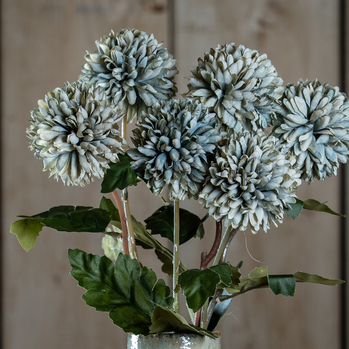 Hill Interiors white chrysanthemum faux stems with layered domed blooms in muted grey-green tones and green leaves, arranged in a ceramic vase against wood.