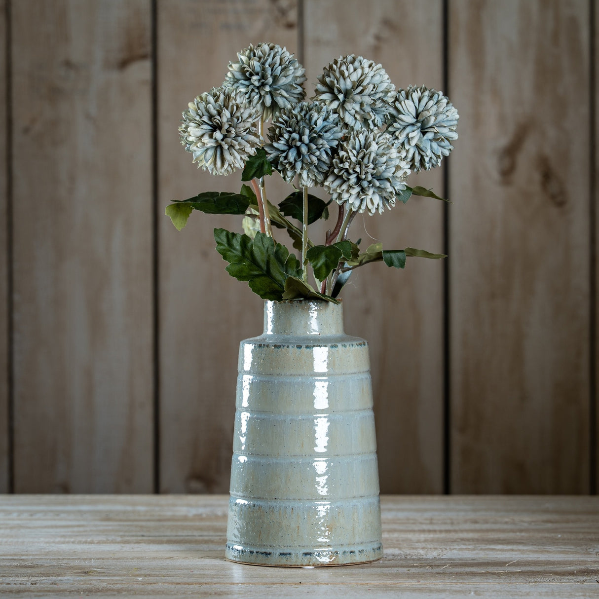 Hill Interiors White Chrysanthemum faux stems arranged in a glossy pale ceramic ribbed vase on a wooden surface against a plank wall.