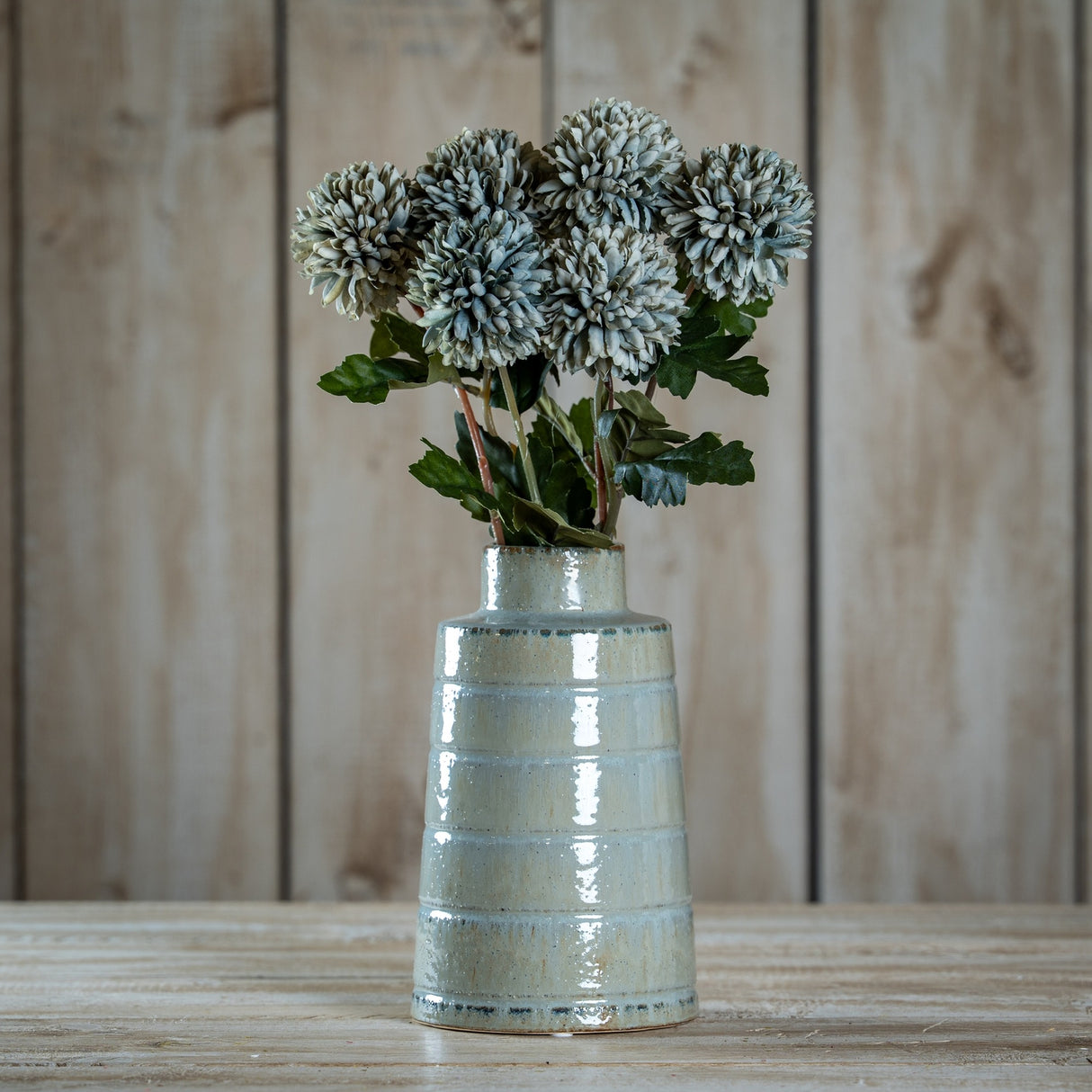 Hill Interiors Pale Green Blue Short Chrysanthemum faux stems arranged in a glossy pale ceramic ribbed vase on a wooden surface against timber panelling.