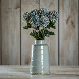 Hill Interiors Pale Green Blue Short Chrysanthemum faux stems arranged in a glossy pale ceramic ribbed vase on a wooden surface against timber panelling.