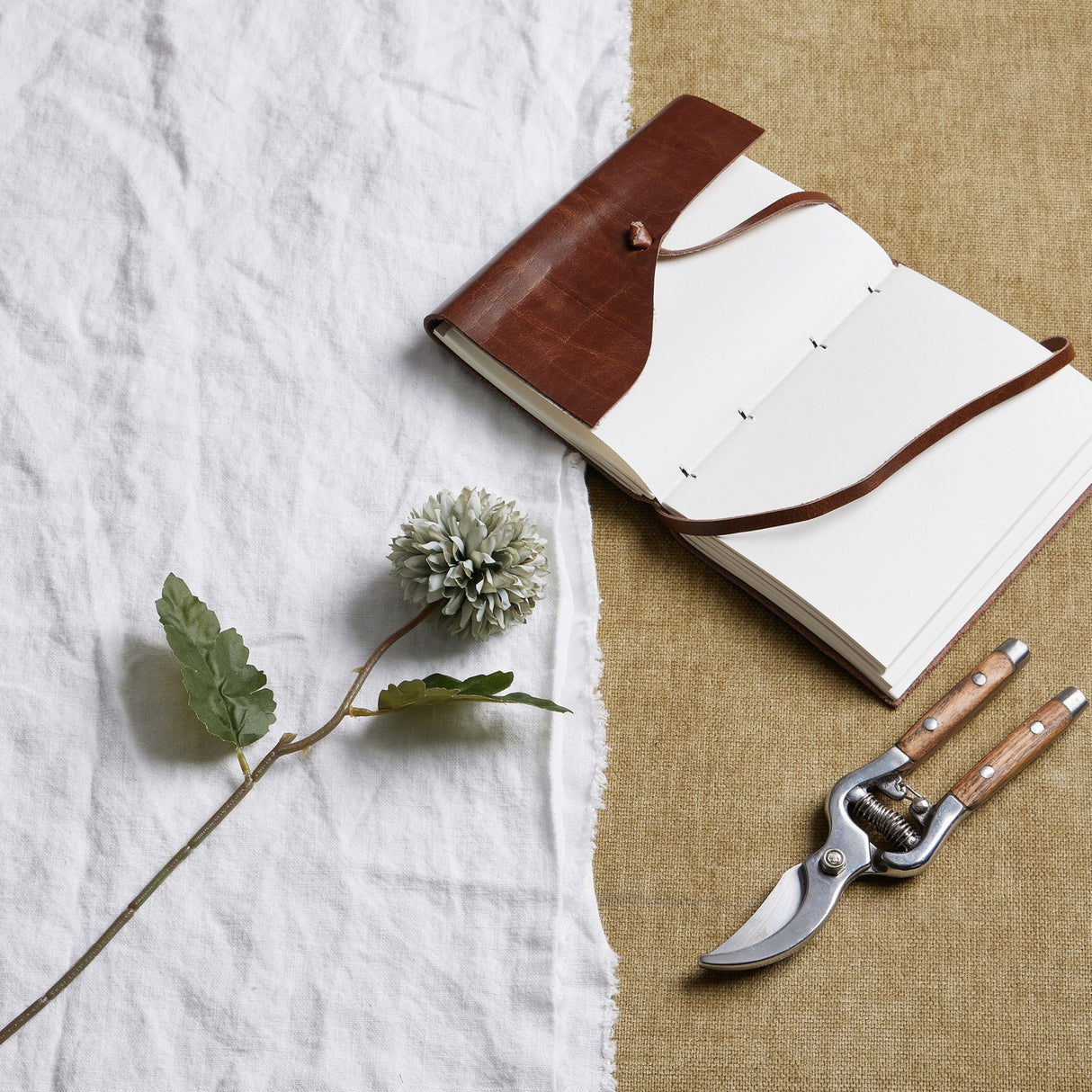 Hill Interiors Pale Green Blue Short Chrysanthemum faux stem laid on white linen beside an open leather journal and wooden-handled secateurs.