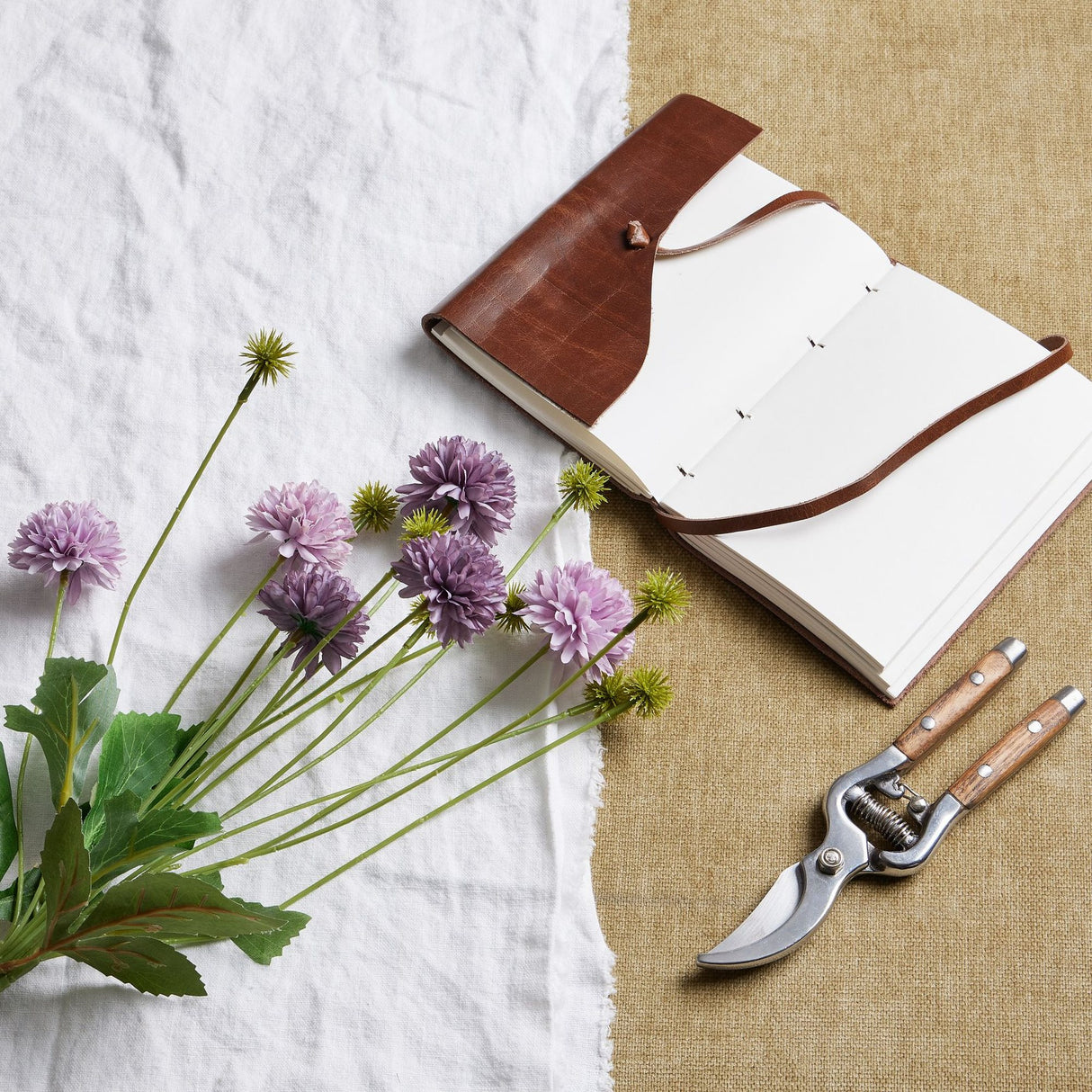 Hill Interiors Purple Chrysanthemum Bouquet with faux pompom blooms and green leaves beside a brown leather journal and wooden-handled secateurs.