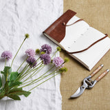 Hill Interiors Purple Chrysanthemum Bouquet with faux pompom blooms and green leaves beside a brown leather journal and wooden-handled secateurs.