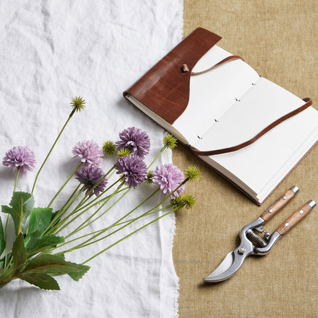 Hill Interiors Purple Chrysanthemum Bouquet with faux pompom blooms and green leaves beside a brown leather journal and wooden-handled secateurs.