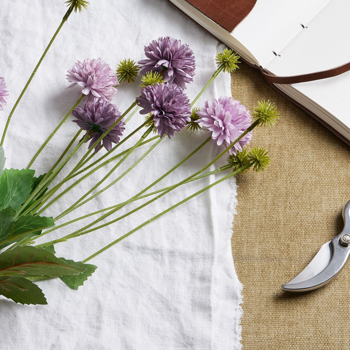 Hill Interiors Purple Chrysanthemum Bouquet with layered pompom heads and green leaves, styled on white linen beside an open leather journal and secateurs.