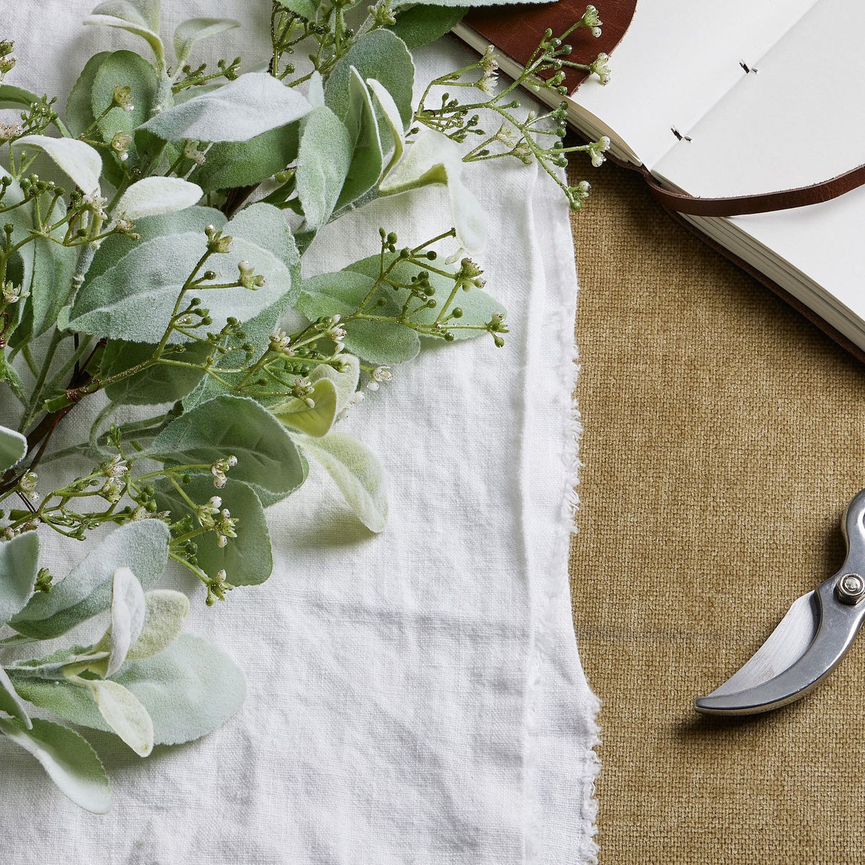 Hill Interiors large winter sprig with velvety lambs ear leaves and fine wax flower sprays on a white linen cloth beside an open leather journal and secateurs.