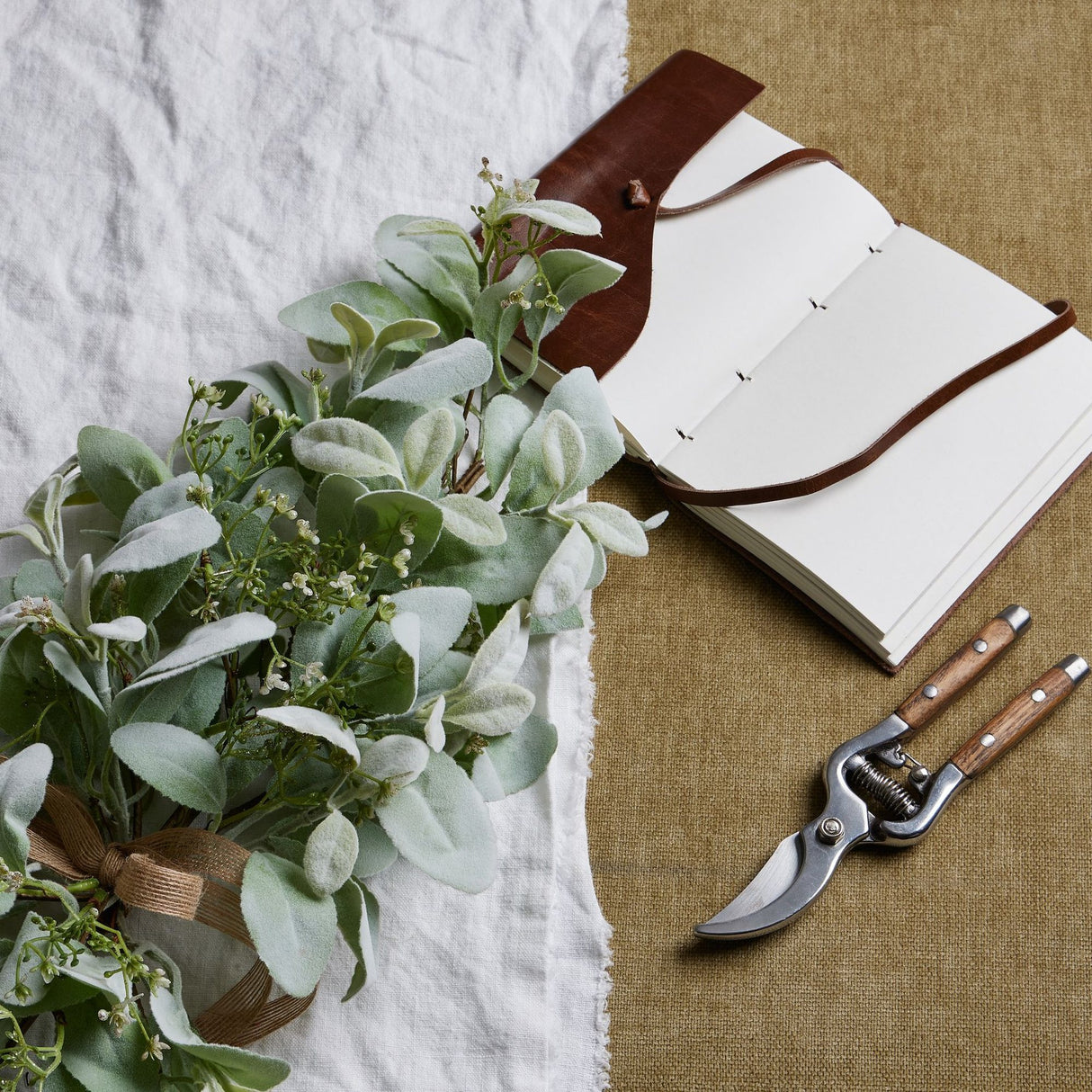 Winter spray arrangement with lambs ear leaves and wax flower sprigs, tied with hessian ribbon beside a leather journal and wooden-handled secateurs.