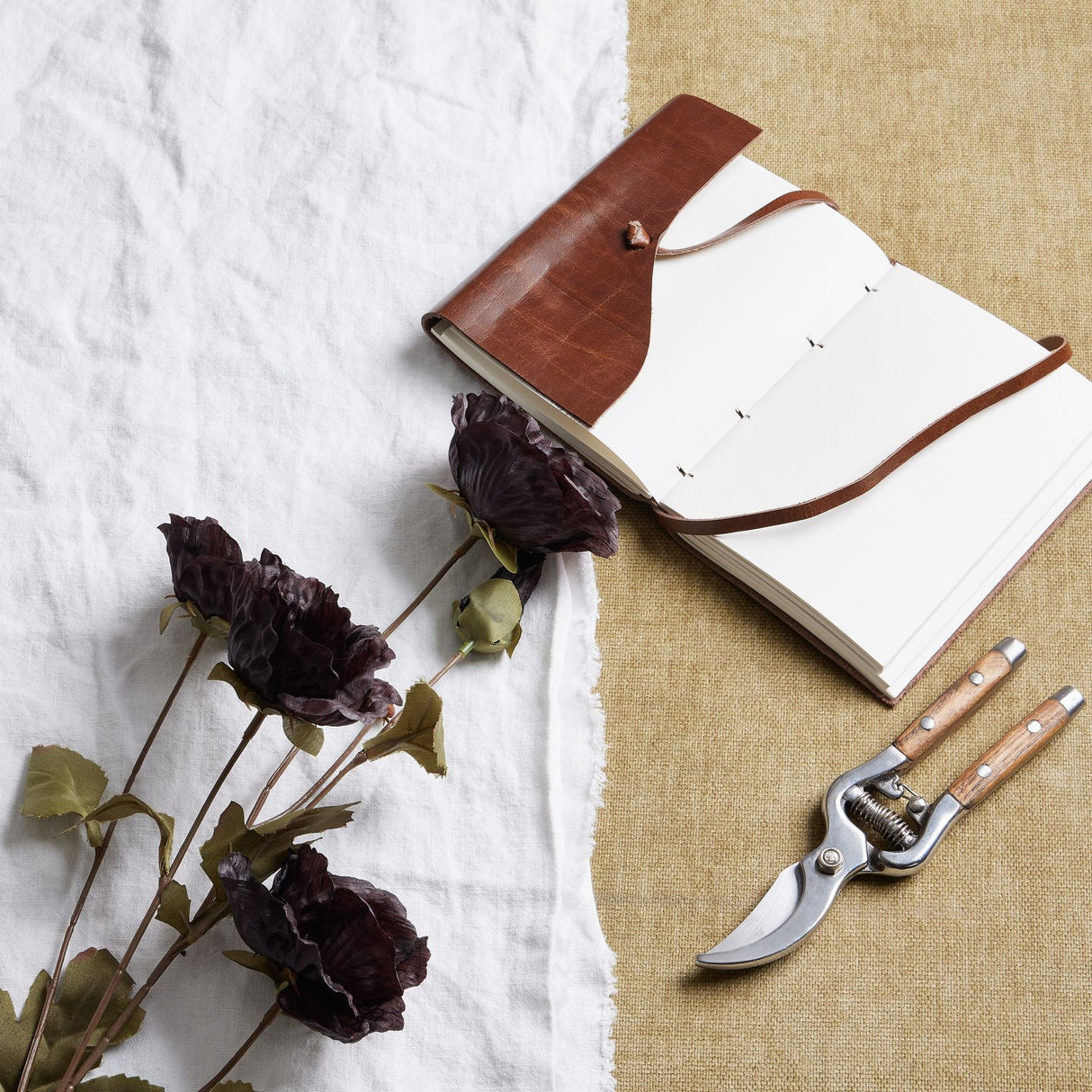 Hill Interiors Tall Black Poppy Stems laid on white linen beside an open leather-bound journal and wooden-handled secateurs on hessian.