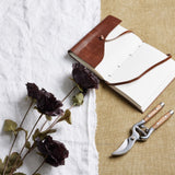 Hill Interiors Tall Black Poppy Stems laid on white linen beside an open leather-bound journal and wooden-handled secateurs on hessian.