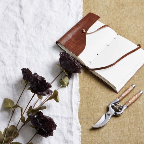 Hill Interiors Tall Black Poppy Stems laid on white linen beside an open leather-bound journal and wooden-handled secateurs on hessian.