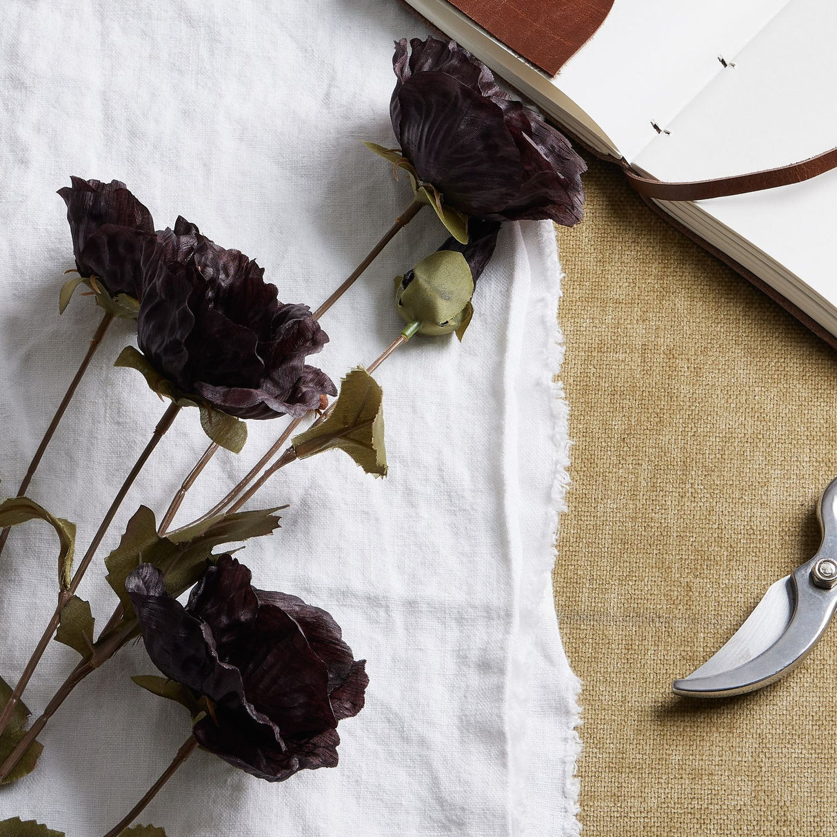 Hill Interiors Tall Black Poppy Stem faux flowers with crinkled petals and green leaves on white linen beside an open leather journal and pruning shears.