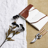 Hill Interiors Black Chrysanthemum faux stem with layered petals and green leaves, laid on white linen beside a leather journal and wooden-handled secateurs.