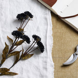 Hill Interiors Black Chrysanthemum faux stem with layered dark blooms and mottled leaves, styled on white linen beside an open leather journal and secateurs.