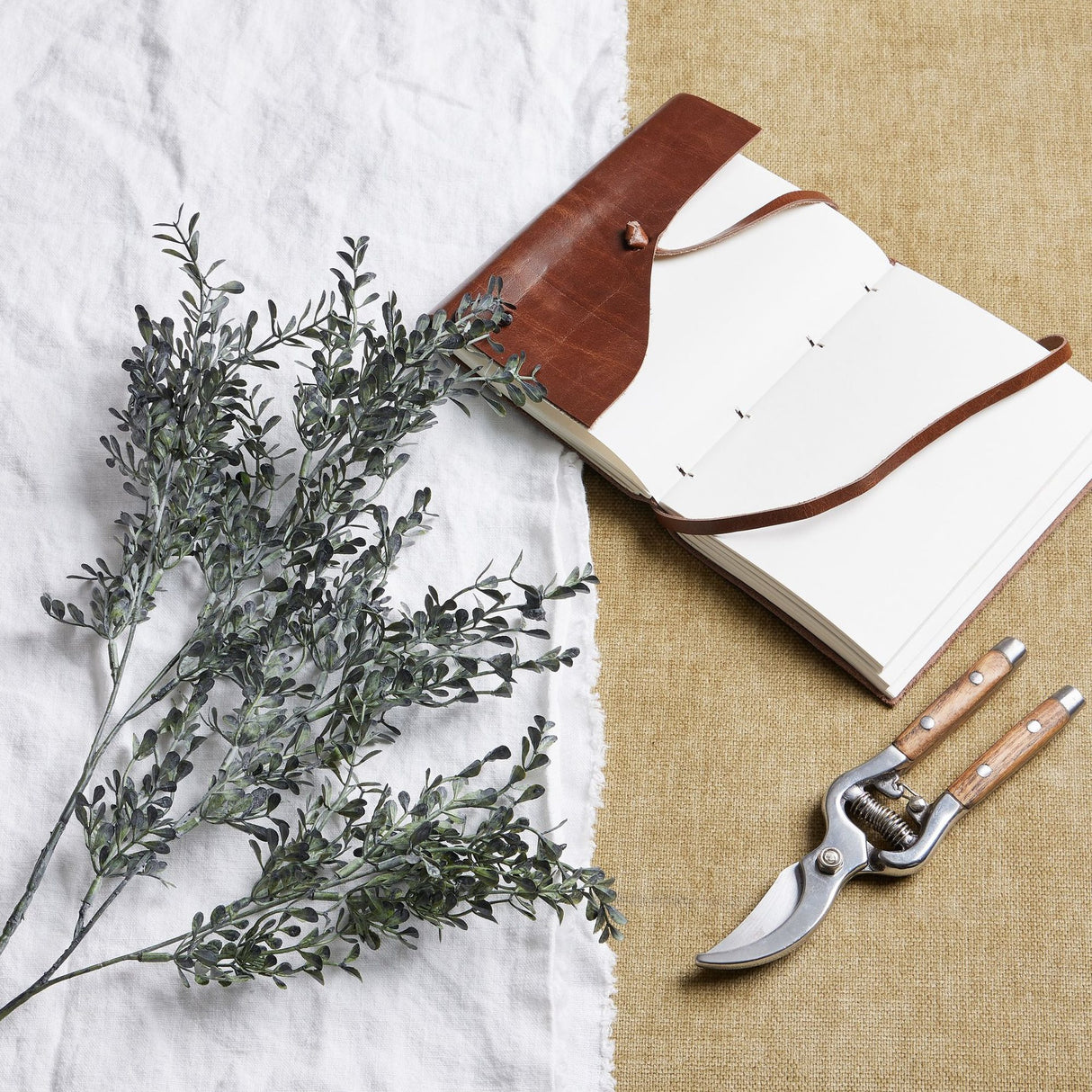 Hill Interiors Blackened Grass Spray Stem laid on white linen beside a leather-bound journal and wooden-handled secateurs on hessian.