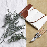 Hill Interiors Blackened Grass Spray Stem laid on white linen beside a leather-bound journal and wooden-handled secateurs on hessian.